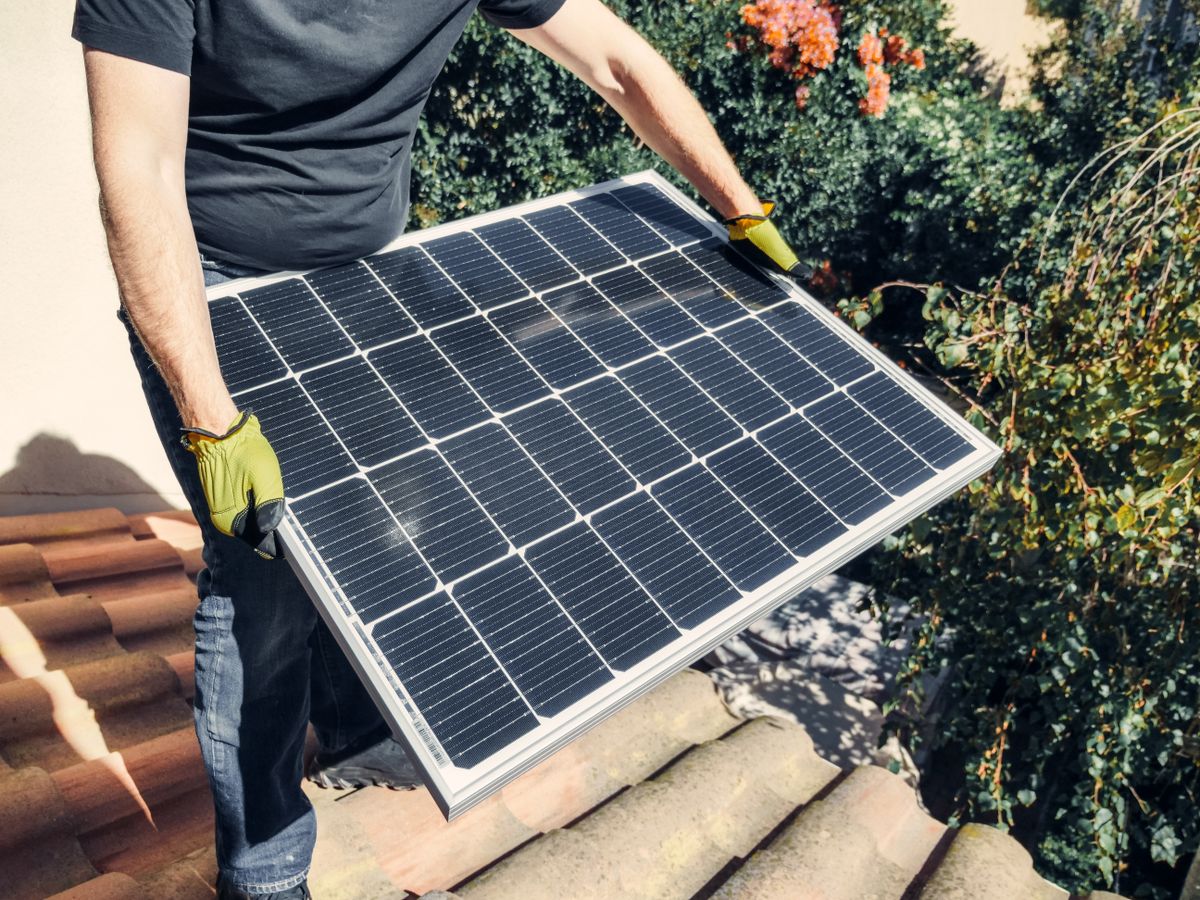 Worker carrying a large solar panel across a tile rooftop